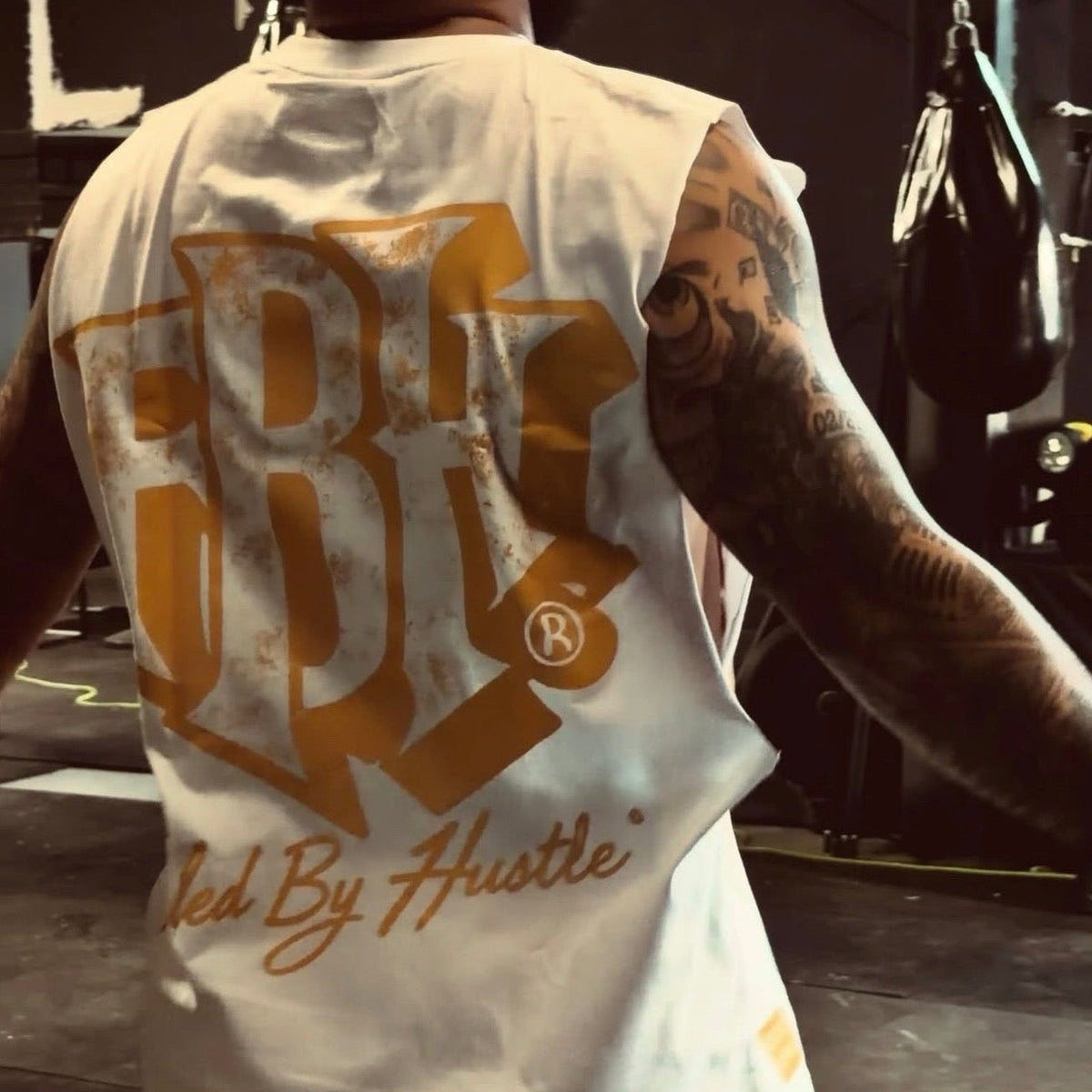 A person viewed from behind wearing a sleeveless white tank top with a large distressed gold graphic on the back and the words 'Fed By Hustle' in gold script. The setting appears to be a dim gym with punching bags in the background.