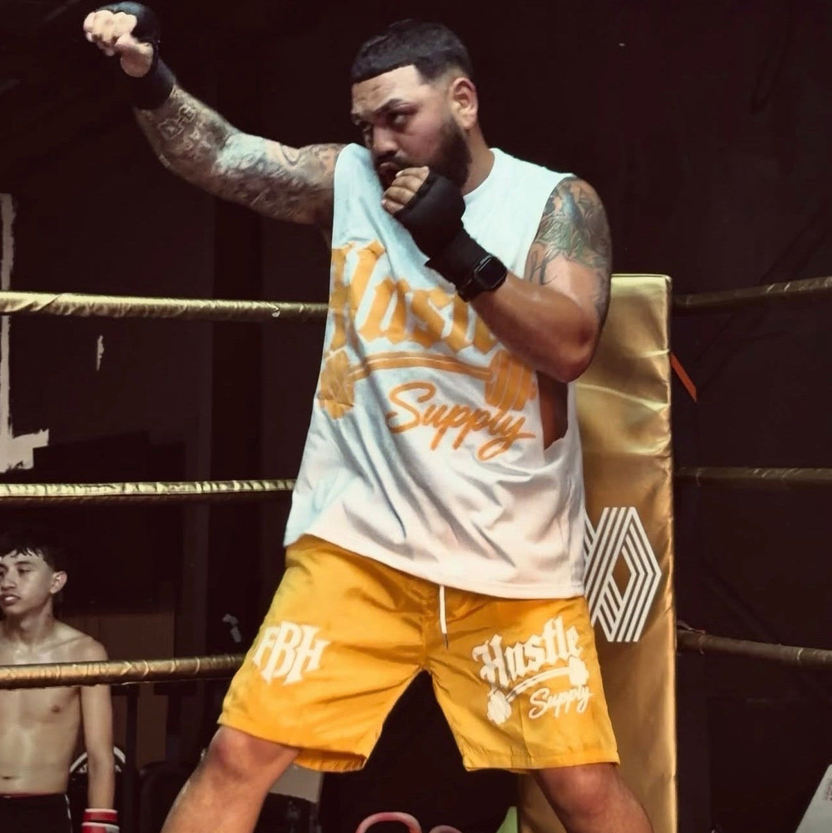 A man in a boxing ring wearing a white sleeveless tank top and yellow shorts with 'Hustle Supply' printed on them, posing with one arm raised. Another boxer is visible in the background.